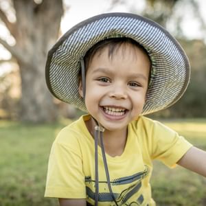Smiling child in a yellow shirt and wide-brim hat outdoors in a park looking at camera