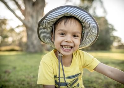 Smiling child in a yellow shirt and wide-brim hat outdoors in a park looking at camera