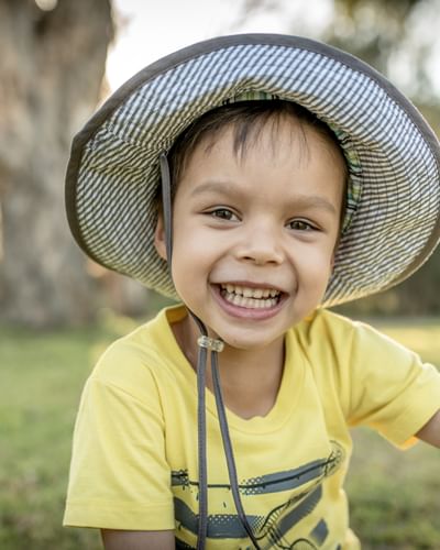 Smiling child in a yellow shirt and wide-brim hat outdoors in a park looking at camera