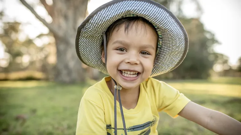 Smiling child in a yellow shirt and wide-brim hat outdoors in a park looking at camera
