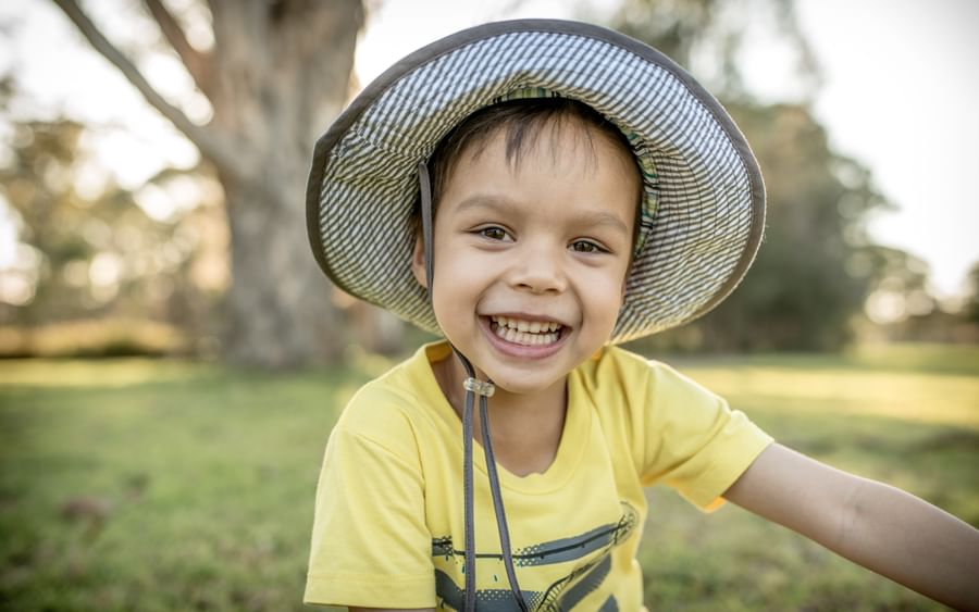 Smiling child in a yellow shirt and wide-brim hat outdoors in a park looking at camera