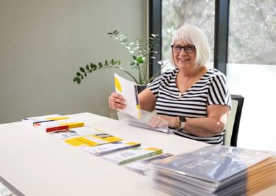 Person with short white hair and glasses smiles while flipping through brochures at a white table.
