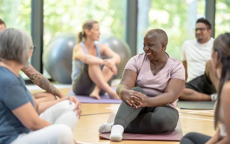Diverse group in a yoga class sits on mats, smiling and chatting.