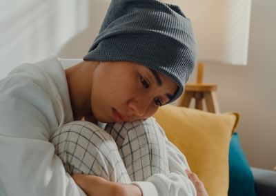 Person wearing a grey beanie and white sweater sits on a couch, hugging knees, looking down.