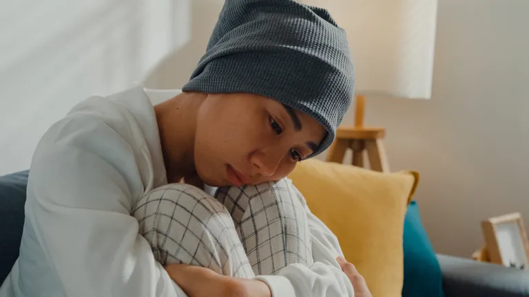 Person wearing a grey beanie and white sweater sits on a couch, hugging knees, looking down.