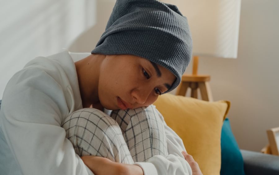 Person wearing a grey beanie and white sweater sits on a couch, hugging knees, looking down.