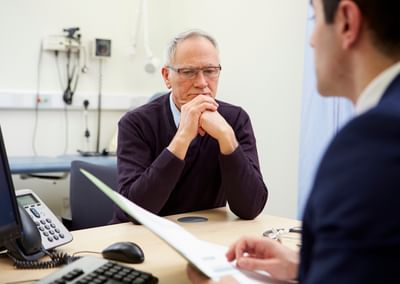 Older person with grey hair and glasses sits at a clinic desk, hands clasped, conversing.