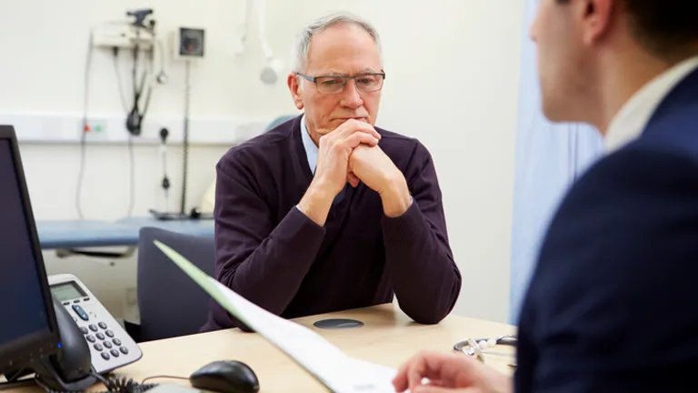 Older person with grey hair and glasses sits at a clinic desk, hands clasped, conversing.