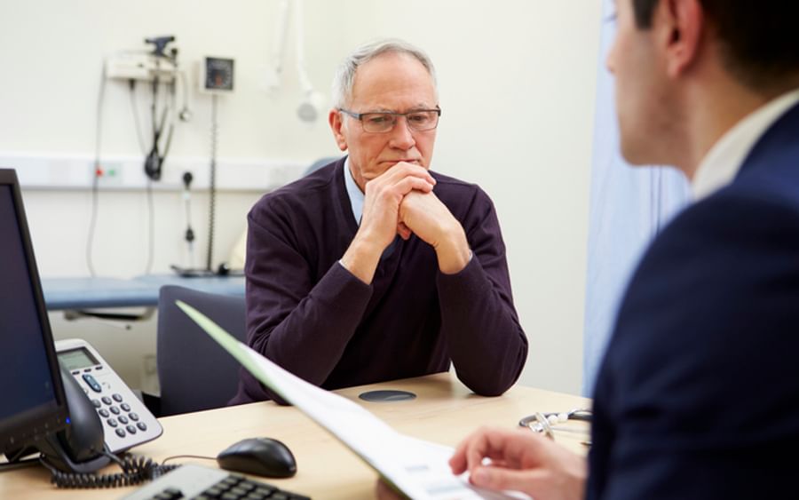 Older person with grey hair and glasses sits at a clinic desk, hands clasped, conversing.