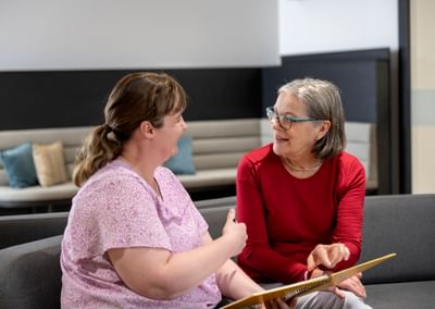 Two people sit on a sofa, chatting; one in a pink top, the other in red, holding a notebook.