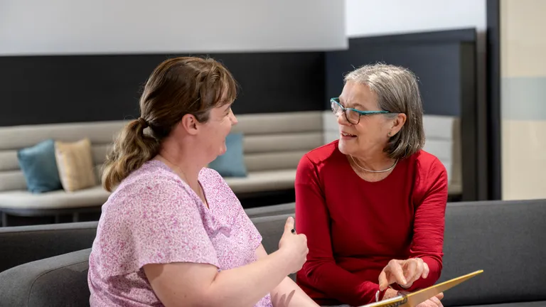 Two people sit on a sofa, chatting; one in a pink top, the other in red, holding a notebook.