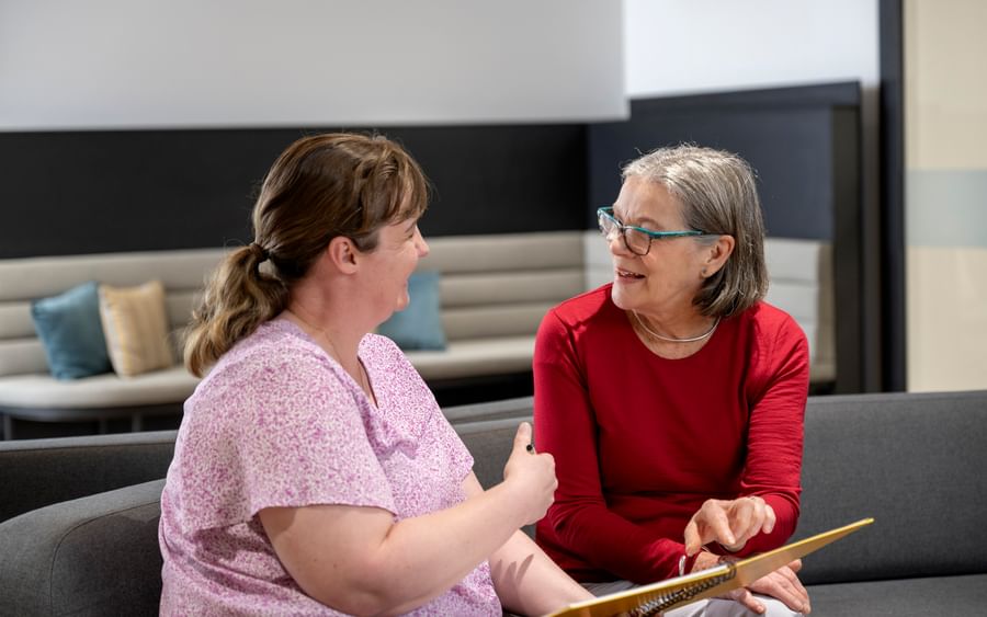 Two people sit on a sofa, chatting; one in a pink top, the other in red, holding a notebook.