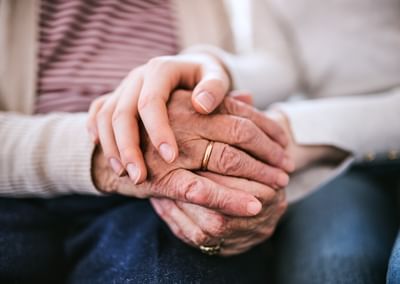 Two hands clasped: an elderly hand with a wedding ring held by a younger hand.