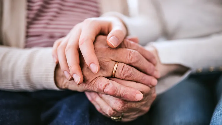 Two hands clasped: an elderly hand with a wedding ring held by a younger hand.