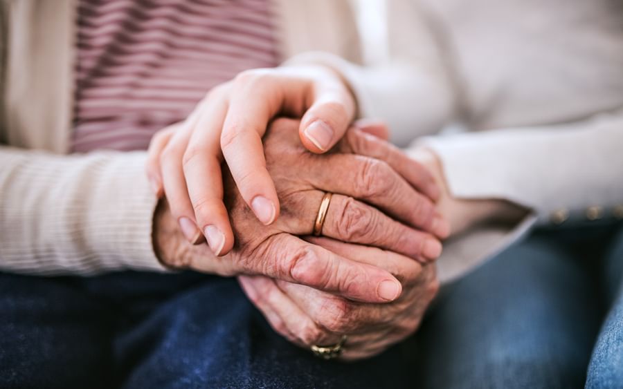 Two hands clasped: an elderly hand with a wedding ring held by a younger hand.
