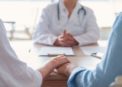 Two people hold hands across a table while a doctor in a white coat sits blurred in the background.