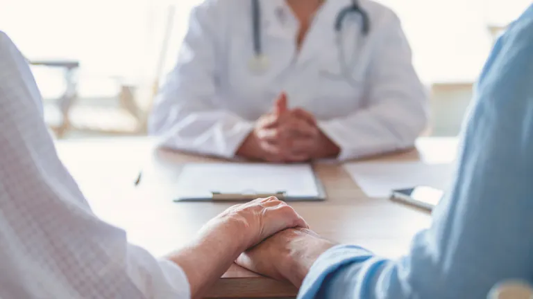 Two people hold hands across a table while a doctor in a white coat sits blurred in the background.