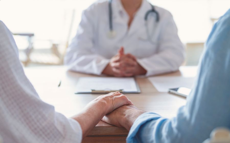 Two people hold hands across a table while a doctor in a white coat sits blurred in the background.
