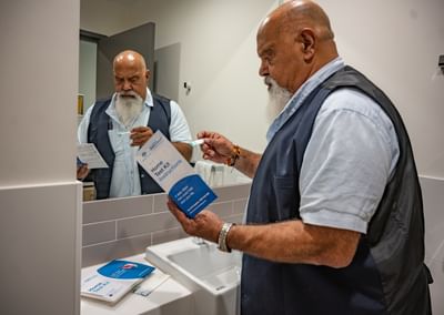 Bearded person in a blue vest reads home test kit instructions while holding a small item near sink.