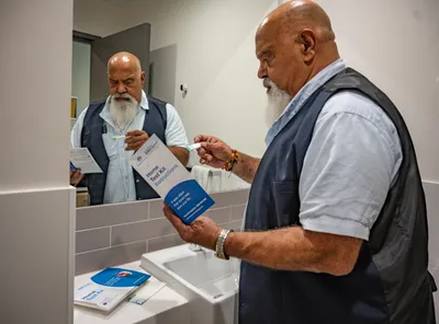 Bearded person in a blue vest reads home test kit instructions while holding a small item near sink.