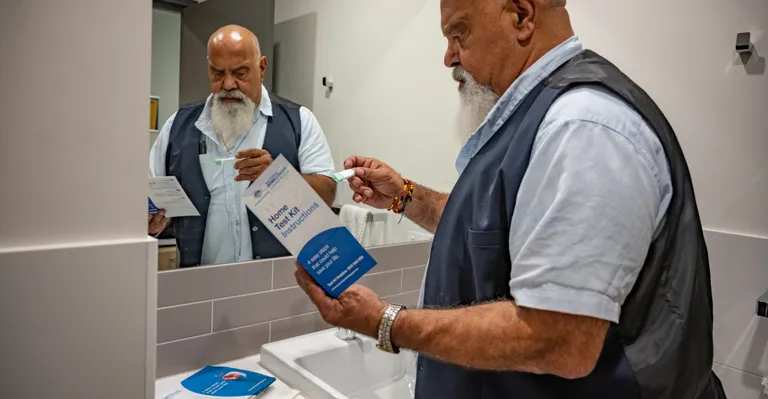 Bearded person in a blue vest reads home test kit instructions while holding a small item near sink.