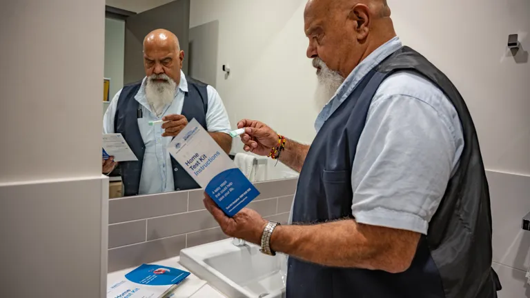 Bearded person in a blue vest reads home test kit instructions while holding a small item near sink.