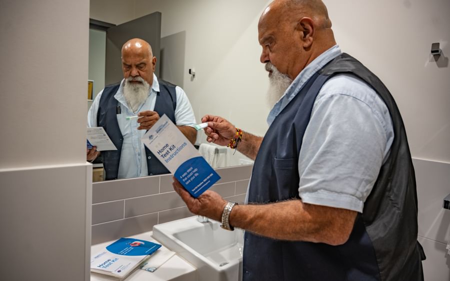 Bearded person in a blue vest reads home test kit instructions while holding a small item near sink.