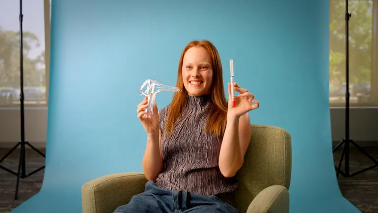 Person with red hair sits in chair against a blue backdrop holding a clear dental retainer and tube