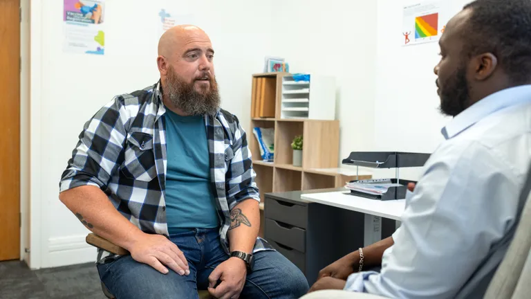 Bearded person in teal shirt and plaid shirt sits across from a clinician in a bright office.