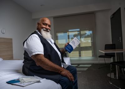 Bearded person sitting on a bed, smiling and holding a brochure in a bright room with window blinds.