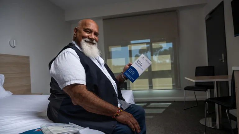 Bearded person sitting on a bed, smiling and holding a brochure in a bright room with window blinds.