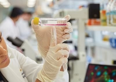 Two lab researchers in white coats and gloves inspect a small vial with a yellow cap.