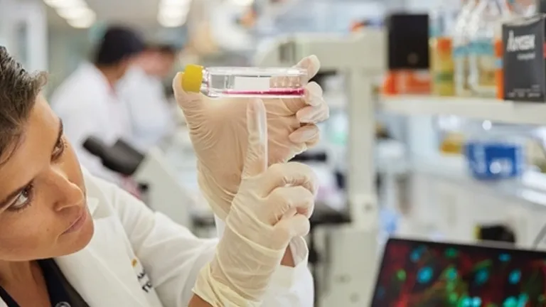 Two lab researchers in white coats and gloves inspect a small vial with a yellow cap.