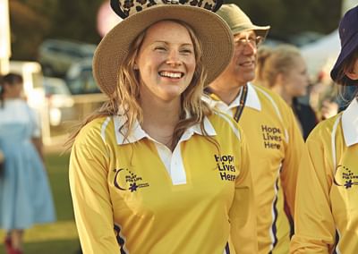 Smiling person in a yellow polo with Hope Lives Here, wearing a large straw hat at an outdoor event.