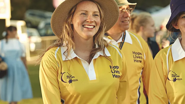 Smiling person in a yellow polo with Hope Lives Here, wearing a large straw hat at an outdoor event.