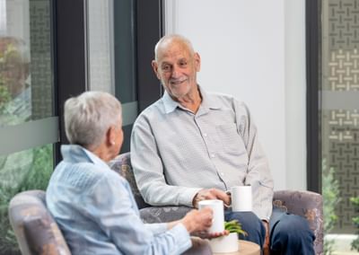 Two older adults chat in a bright, modern lounge by large windows, holding mugs.