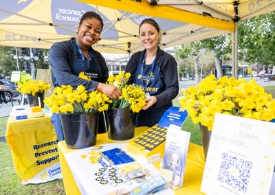 Two smiling volunteers in blue aprons at a Cancer Council stall with yellow flowers outdoors.