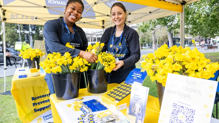 Two smiling volunteers in blue aprons at a Cancer Council stall with yellow flowers outdoors.