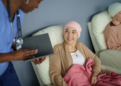 Healthcare worker in blue scrubs shows a clipboard to a patient with a pink headscarf and pink blanket.