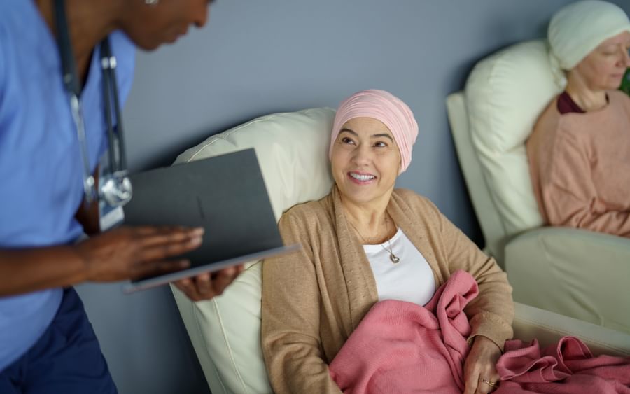 Healthcare worker in blue scrubs shows a clipboard to a patient with a pink headscarf and pink blanket.