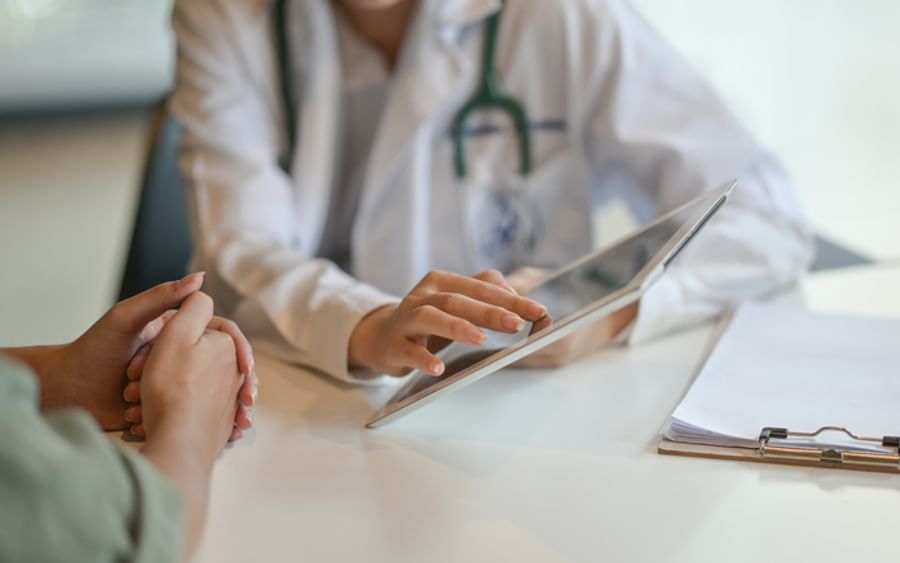 Healthcare professional in a white coat with a stethoscope uses a tablet during patient consultation.
