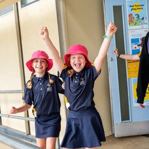 Two children in navy polo shirts and pink hats cheer with arms up; adult in dark hoodie nearby.