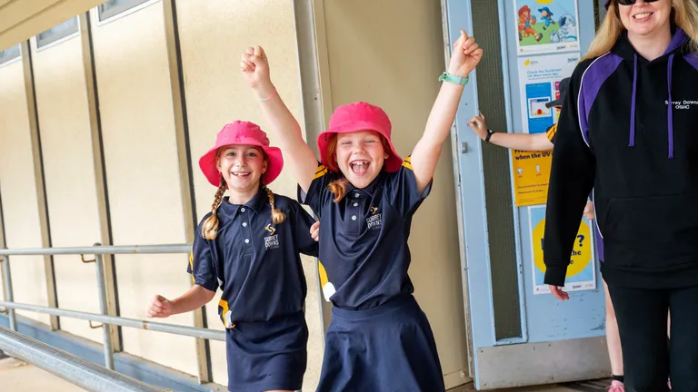Two children in navy polo shirts and pink hats cheer with arms up; adult in dark hoodie nearby.