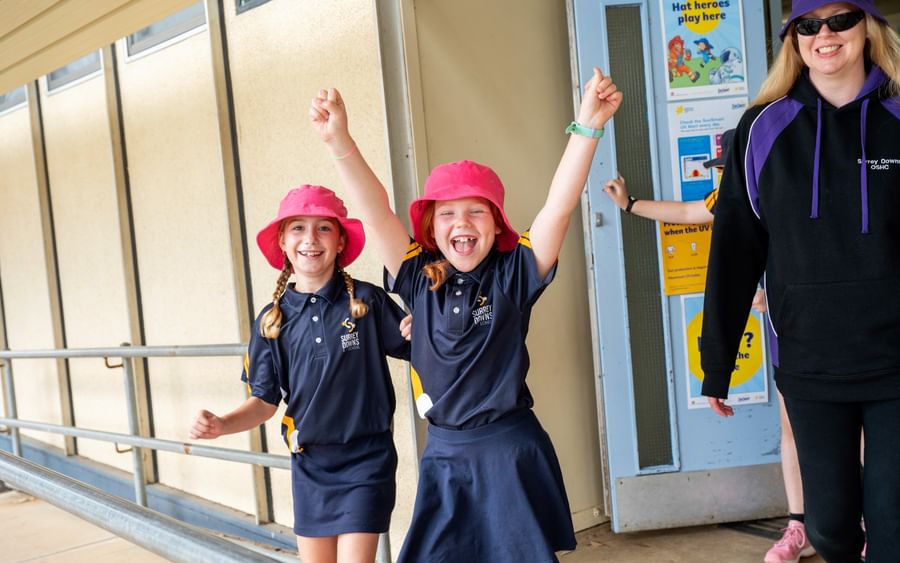 Two children in navy polo shirts and pink hats cheer with arms up; adult in dark hoodie nearby.