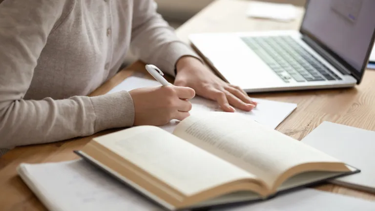 Person writing in a notebook at a wooden desk with an open book and a laptop nearby.
