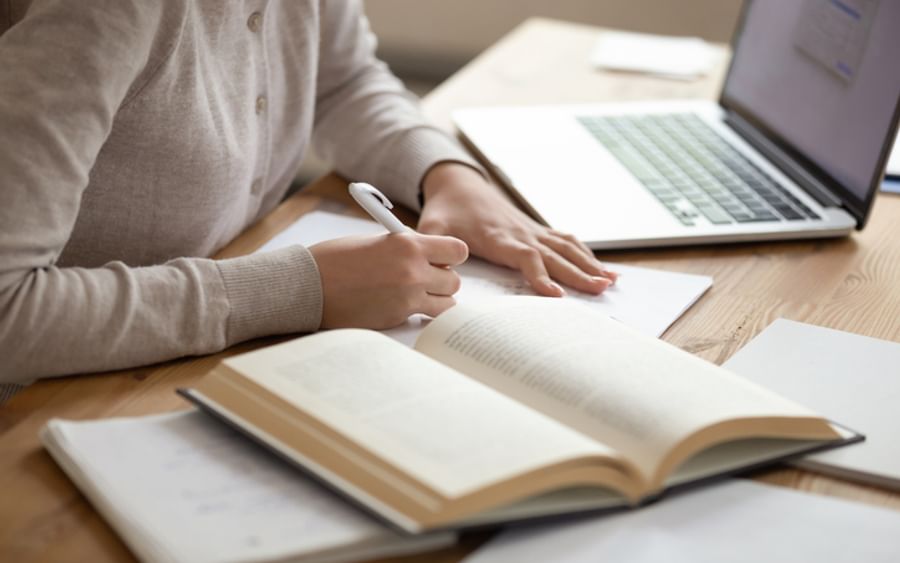Person writing in a notebook at a wooden desk with an open book and a laptop nearby.