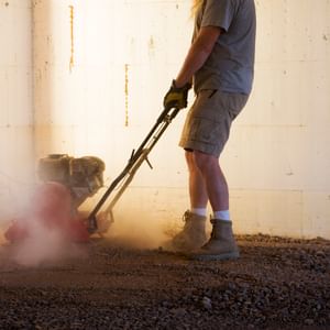 Person wearing gloves and boots, operating a plate compactor on dusty ground indoors.