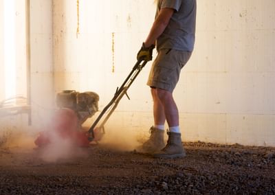 Person wearing gloves and boots, operating a plate compactor on dusty ground indoors.