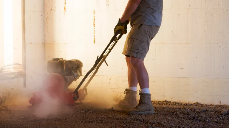 Person wearing gloves and boots, operating a plate compactor on dusty ground indoors.