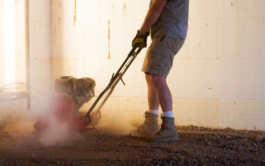 Person wearing gloves and boots, operating a plate compactor on dusty ground indoors.
